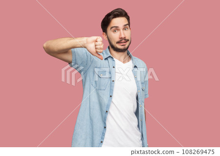 Portrait of bearded man in blue casual style shirt standing assess project, shows sign of dislike, looks with negative expression and disapproval. Indoor studio shot isolated on pink background. 108269475