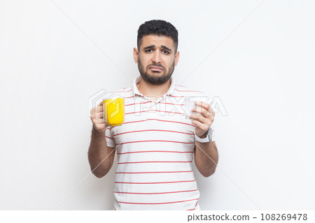 Portrait of sad unhappy unhealthy bearded man wearing striped t-shirt standing with yellow cup, looking at camera, feels bad, frowning face. Indoor studio shot isolated on gray background. 108269478