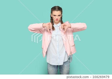 Portrait of displeased unhappy teenager girl with braids wearing pink jacket standing with thumbs down, showing dislike gesture to camera. Indoor studio shot isolated on green background. 108269479