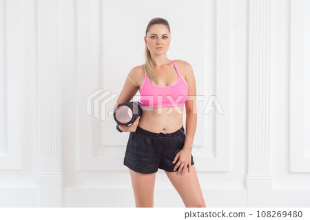 Portrait of confident athletic sporty beautiful blonde woman with perfect body in pink top and black shorts standing, holding foam roll and looking at camera. Indoor studio shot. 108269480