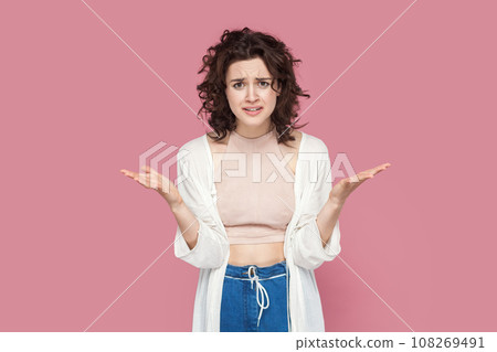 Portrait of angry attractive young adult woman with curly hair wearing casual style outfit raised her hands, asking what, arguing, frowning face. Indoor studio shot isolated on pink background. 108269491