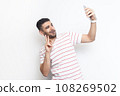 Portrait of joyful handsome playful bearded man wearing striped t-shirt standing making selfie, showing v sign to his followers. Indoor studio shot isolated on gray background. 108269502
