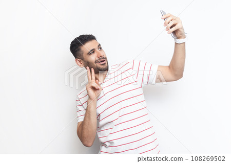 Portrait of joyful handsome playful bearded man wearing striped t-shirt standing making selfie, showing v sign to his followers. Indoor studio shot isolated on gray background. Portrait of joyful handsome playful bearded man wearing striped t-shirt standing making selfie, showing v sign to his followers. Indoor studio shot isolated on gray background. 108269502
