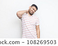 Portrait of handsome sick tired bearded man wearing striped t-shirt standing massaging his painful neck, frowning face from terrible pain. Indoor studio shot isolated on gray background. 108269503