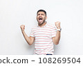 Portrait of extremely happy joyful cheerful bearded man wearing striped t-shirt standing with clenched raised fists, screaming, celebrating victory. Indoor studio shot isolated on gray background. 108269504
