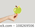 Closeup of woman hand showing paper green apple, symbol of healthy eating. Indoor studio shot isolated on gray background. 108269506