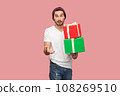 Portrait of puzzled confused bearded man in white T-shirt and beany hat holding two present boxes in hands, looking at camera with surprised expression. Indoor studio shot isolated on pink background 108269510