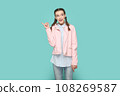 Portrait of excited smiling positive teenager girl with braids wearing pink jacket pointing index finger away, showing copy space for advertisement. Indoor studio shot isolated on green background. 108269587