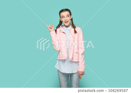 Portrait of excited smiling positive teenager girl with braids wearing pink jacket pointing index finger away, showing copy space for advertisement. Indoor studio shot isolated on green background. Portrait of excited smiling positive teenager girl with braids wearing pink jacket pointing index finger away, showing copy space for advertisement. Indoor studio shot isolated on green background. 108269587
