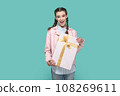 Portrait of excited positive teenager girl wearing pink jacket standing wrapped present box in hands, looking at camera, waiting holiday to give gift. Indoor studio shot isolated on green background. 108269611