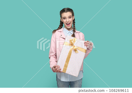 Portrait of excited positive teenager girl wearing pink jacket standing wrapped present box in hands, looking at camera, waiting holiday to give gift. Indoor studio shot isolated on green background. Portrait of excited positive teenager girl wearing pink jacket standing wrapped present box in hands, looking at camera, waiting holiday to give gift. Indoor studio shot isolated on green background. 108269611