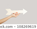Closeup of woman hand holding white arrow pointing away, showing direction aside. Indoor studio shot isolated on gray background. 108269619