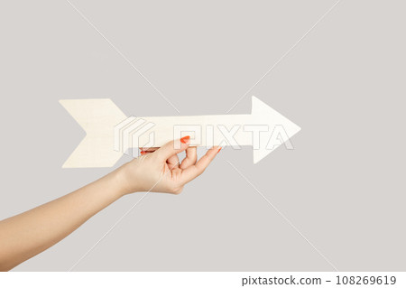 Closeup of woman hand holding white arrow pointing away, showing direction aside. Indoor studio shot isolated on gray background. Closeup of woman hand holding white arrow pointing away, showing direction aside. Indoor studio shot isolated on gray background. 108269619