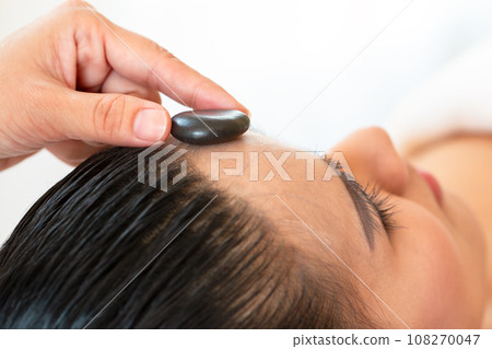 a close up therapist hand palcing black spa stone on customer forehead laying down on bed 108270047
