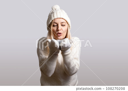 Woman blows on snowflakes in her hands, dressed in mittens, hat and sweater 108270208