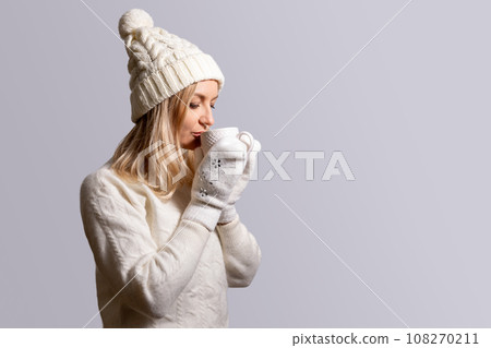 Woman in white sweater, cap with bubo and mittens drinks from cup Woman in white sweater, cap with bubo and mittens drinks from cup 108270211