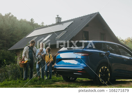 Family with electric car standing in front their house with solar panels on roof. Solar energy and sustainable lifestyle of young family. Concept of green energy and sustainable future for next 108271879