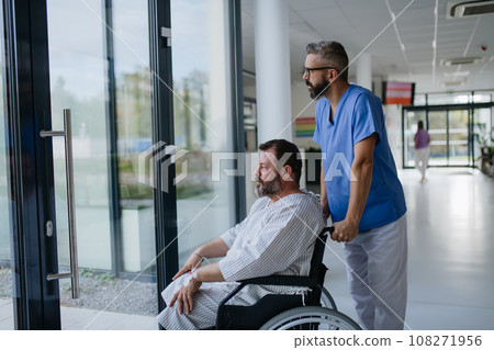 Male nurse pushing a patient in a wheelchair along a hospital corridor. Overweight patient feeling anxious and has health concerns. Male nurse pushing a patient in a wheelchair along a hospital corridor. Overweight patient feeling anxious and has health concerns. 108271956