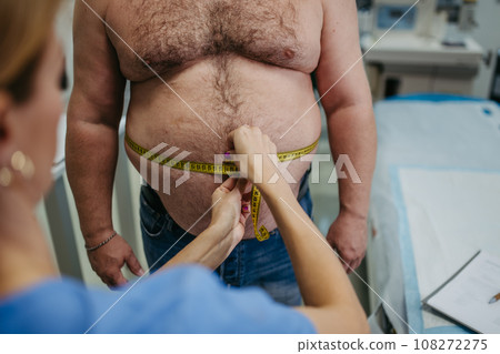 Female doctor measuring waist of overweight patient using tape measure. Obesity affecting middle-aged men's health. Concept of health risks of overwight and obesity. 108272275