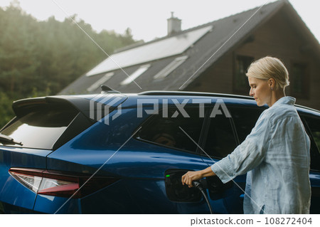 Portrait of a woman charging electric car in front of her house with solar panels on roof. Mother plugging the charger into the charging port. Solar energy and sustainable lifestyle of young family. Portrait of a woman charging electric car in front of her house with solar panels on roof. Mother plugging the charger into the charging port. Solar energy and sustainable lifestyle of young family. 108272404