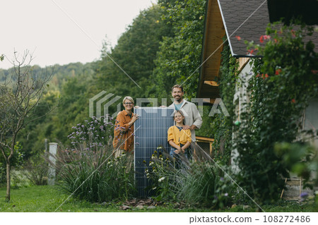 Father mother and daughter standing in garden with solar panel. Solar energy and sustainable lifestyle of young family. Concept of green energy and sustainable future for next generations. Father mother and daughter standing in garden with solar panel. Solar energy and sustainable lifestyle of young family. Concept of green energy and sustainable future for next generations. 108272486