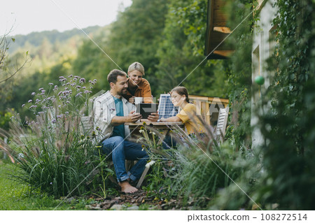 Parents with daughter sitting outdoors in garden, with model of solar panel, learning about solar energy. Solar energy and sustainable lifestyle of young family. Concept of green energy and 108272514