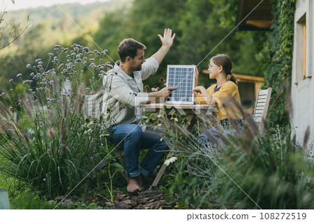 Father with daughter sitting outdoors in garden, with model of solar panel. Solar energy and sustainable lifestyle of young family. Concept of green energy and sustainable future for next generations. Father with daughter sitting outdoors in garden, with model of solar panel. Solar energy and sustainable lifestyle of young family. Concept of green energy and sustainable future for next generations. 108272519