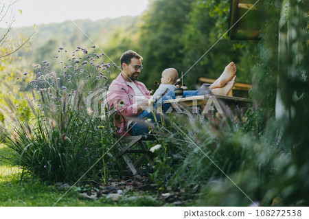 Man sitting in the garden with feet on table, playing with his baby son. Father having bonding moment with his little kid. Man sitting in the garden with feet on table, playing with his baby son. Father having bonding moment with his little kid. 108272538