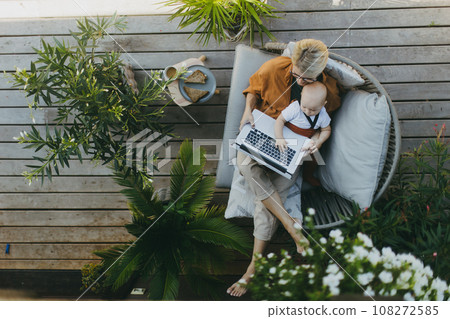 Top view of mother holding baby while working on laptop in garden. Businesswoman working remotely from outdoor home office and taking care of little son. Life work balance with kid. 108272585