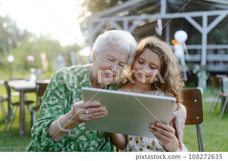 Young girl showing someting on tablet to elderly grandmother at garden party. Love and closeness between grandparent and grandchild. 108272635