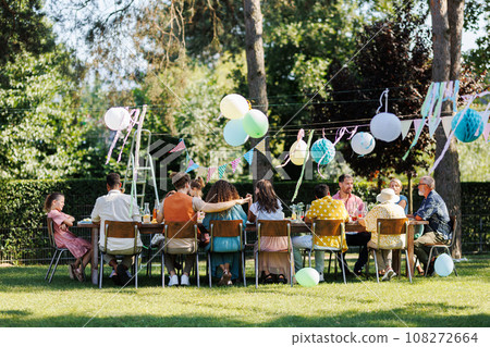 Family and friends sitting at the party table during a summer garden party outdoors. Rear view of people sitting at the table. Family and friends sitting at the party table during a summer garden party outdoors. Rear view of people sitting at the table. 108272664
