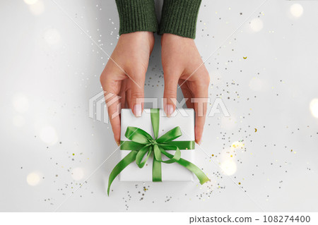 Hands holding Xmas gift package in white wrapping paper with green ribbon on festive background. Zero waste pack Christmas present packaging. Happy holidays concept. Flat lay, top view 108274400