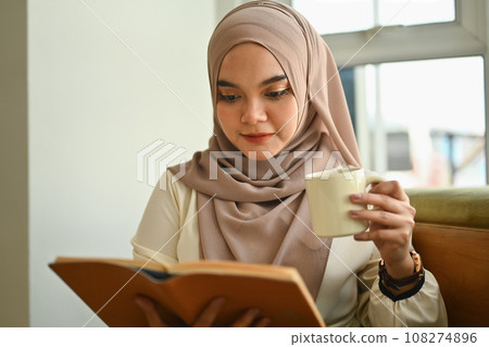 Relaxed muslim woman drinking hot tea and reading book in coffee shop Relaxed muslim woman drinking hot tea and reading book in coffee shop 108274896
