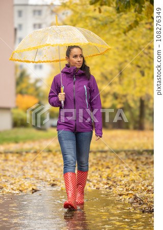 A nice brunette in warm clothes and with an umbrella is walking in a city park surrounded by fallen autumn leaves 108276349