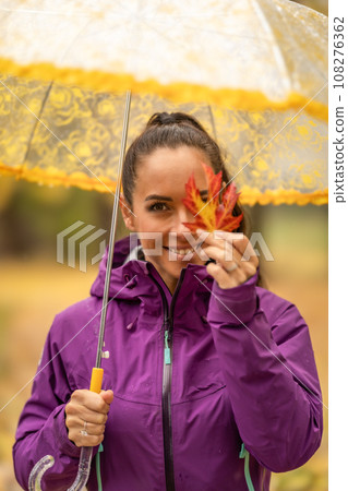 Cute young woman under an umbrella holding a colorful maple leaf and covering her eye in an autumn park 108276362