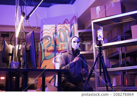 Hacker in anonymous mask threatening online while streaming live in abandoned warehouse. Internet scammer recording video with ransom on smartphone in hideout place at night 108276864