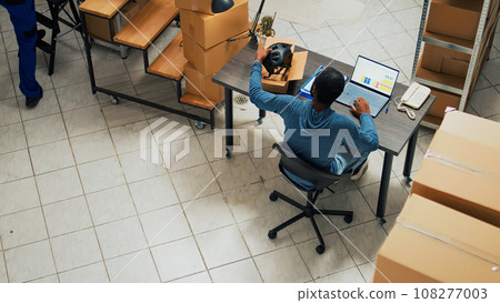 Male startup owner working on financial planning and logistics with laptop, checking merchandise before shipping it. Employee taking notes for stock inventory and sending order. Tripod shot. Male startup owner working on financial planning and logistics with laptop, checking merchandise before shipping it. Employee taking notes for stock inventory and sending order. Tripod shot. 108277003