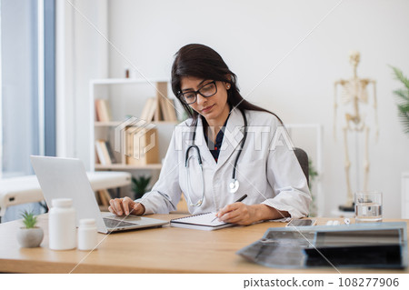 Female doctor with laptop writing prescription at office Female doctor with laptop writing prescription at office 108277906
