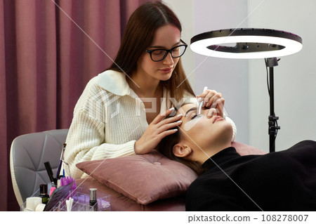 beautician with tweezers makes eyelash extension for young woman in beauty salon. beautician with tweezers makes eyelash extension for young woman in beauty salon. 108278007