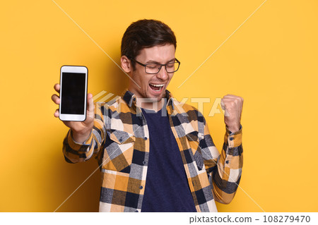 Excited glad young male posing with smartphone over yellow background Excited glad young male posing with smartphone over yellow background 108279470