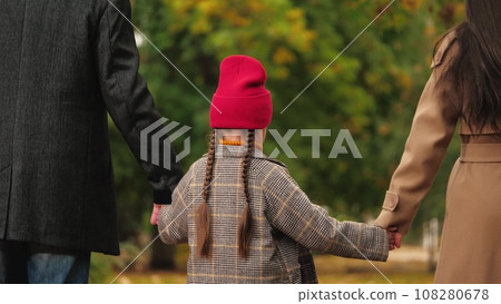 Cute little girl holds mother and father hands walking across autumn park. Happy little daughter spends weekend with parents in park. Mother, father and little daughter rest in fall garden Cute little girl holds mother and father hands walking across autumn park. Happy little daughter spends weekend with parents in park. Mother, father and little daughter rest in fall garden 108280678