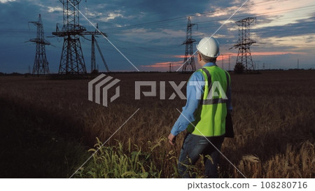 Skilled electrician with helmet and laptop walks across wheat field to power transmission lines at sunset. Technician ready to works with power facility holding laptop in open country field 108280716