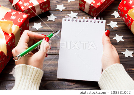 Top view of female hand writing in a notebook on wooden Christmas background. Wish list. New Year concept Top view of female hand writing in a notebook on wooden Christmas background. Wish list. New Year concept 108281062