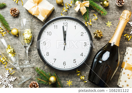 New year celebration concept with a bottle of champagne and two glasses toasting. Christmas gift box, alarm clock and fir tree branch on colored table. Top view Copy space 108281122