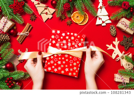 Top view of female hands holding a Christmas present on festive red background. Fir tree and holiday decorations. New Year holiday concept Top view of female hands holding a Christmas present on festive red background. Fir tree and holiday decorations. New Year holiday concept 108281343