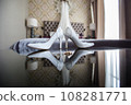 Elegant white bridal heels and wedding ring on a reflective surface, with a luxuriously blurred room backdrop. 108281771