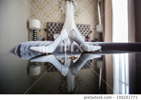 Elegant white bridal heels and wedding ring on a reflective surface, with a luxuriously blurred room backdrop. Elegant white bridal heels and wedding ring on a reflective surface, with a luxuriously blurred room backdrop. 108281771