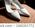 Elegant white bridal heels and wedding ring on a reflective surface, with a luxuriously blurred room backdrop. 108281772