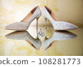 Elegant white bridal heels and wedding ring on a reflective surface, with a luxuriously blurred room backdrop. 108281773