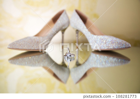 Elegant white bridal heels and wedding ring on a reflective surface, with a luxuriously blurred room backdrop. Elegant white bridal heels and wedding ring on a reflective surface, with a luxuriously blurred room backdrop. 108281773